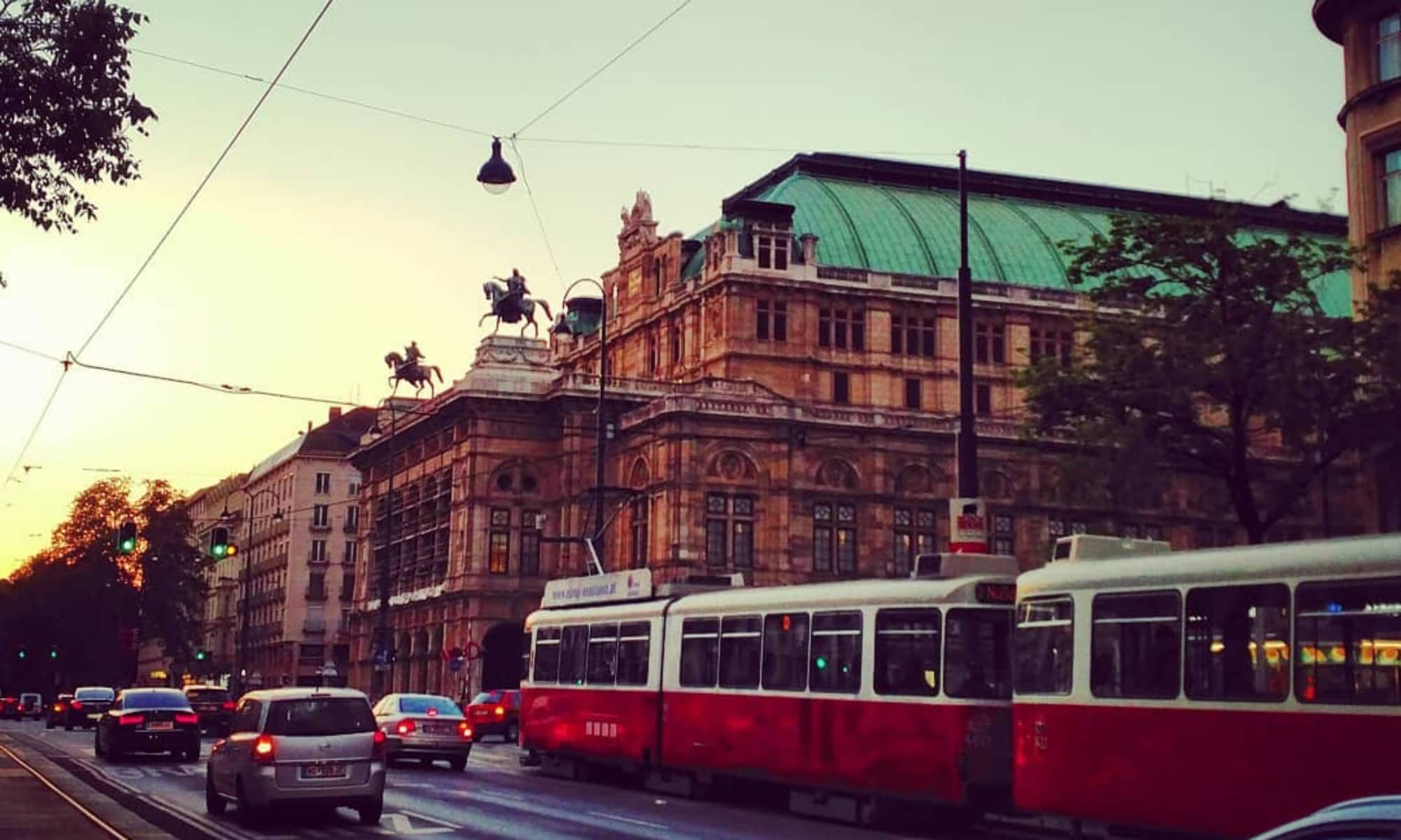 Stra&szlig;enansicht der Wiener Staatsoper bei Sonnenuntergang, mit einer roten Stra&szlig;enbahn und Autos im Vordergrund.