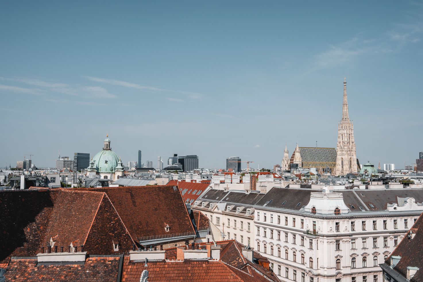 Blick &uuml;ber die D&auml;cher von Wien mit dem Stephansdom und dem gr&uuml;nen Kuppeldach einer Kirche im Vordergrund, moderner Skyline im Hintergrund und klarem Himmel.