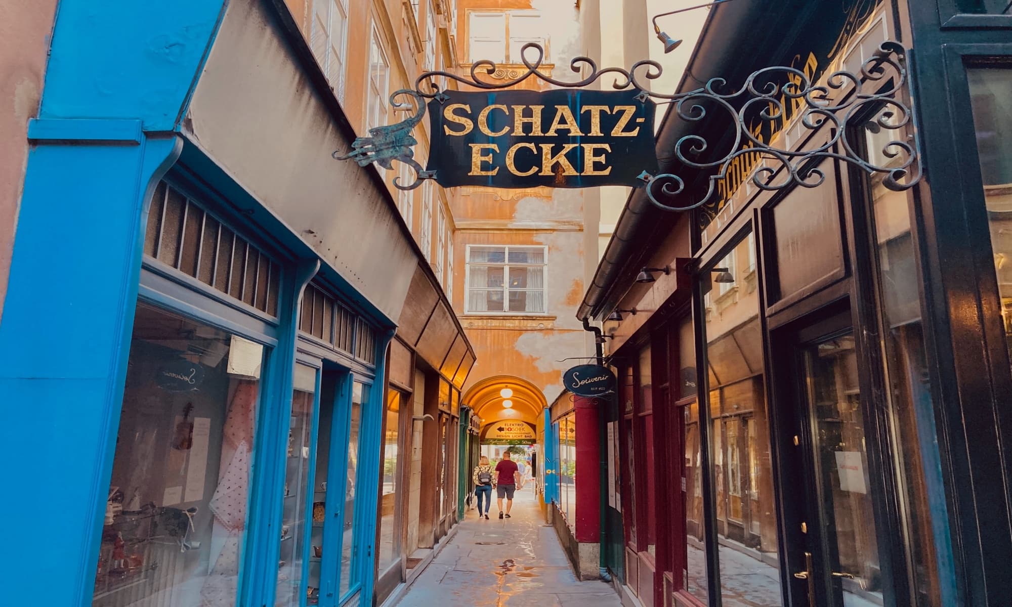 Schmale Gasse in Wien mit dem Schild "Schatz-Ecke" &uuml;ber dem Eingang, flankiert von bunten Ladenfassaden und einer Arkade im Hintergrund.