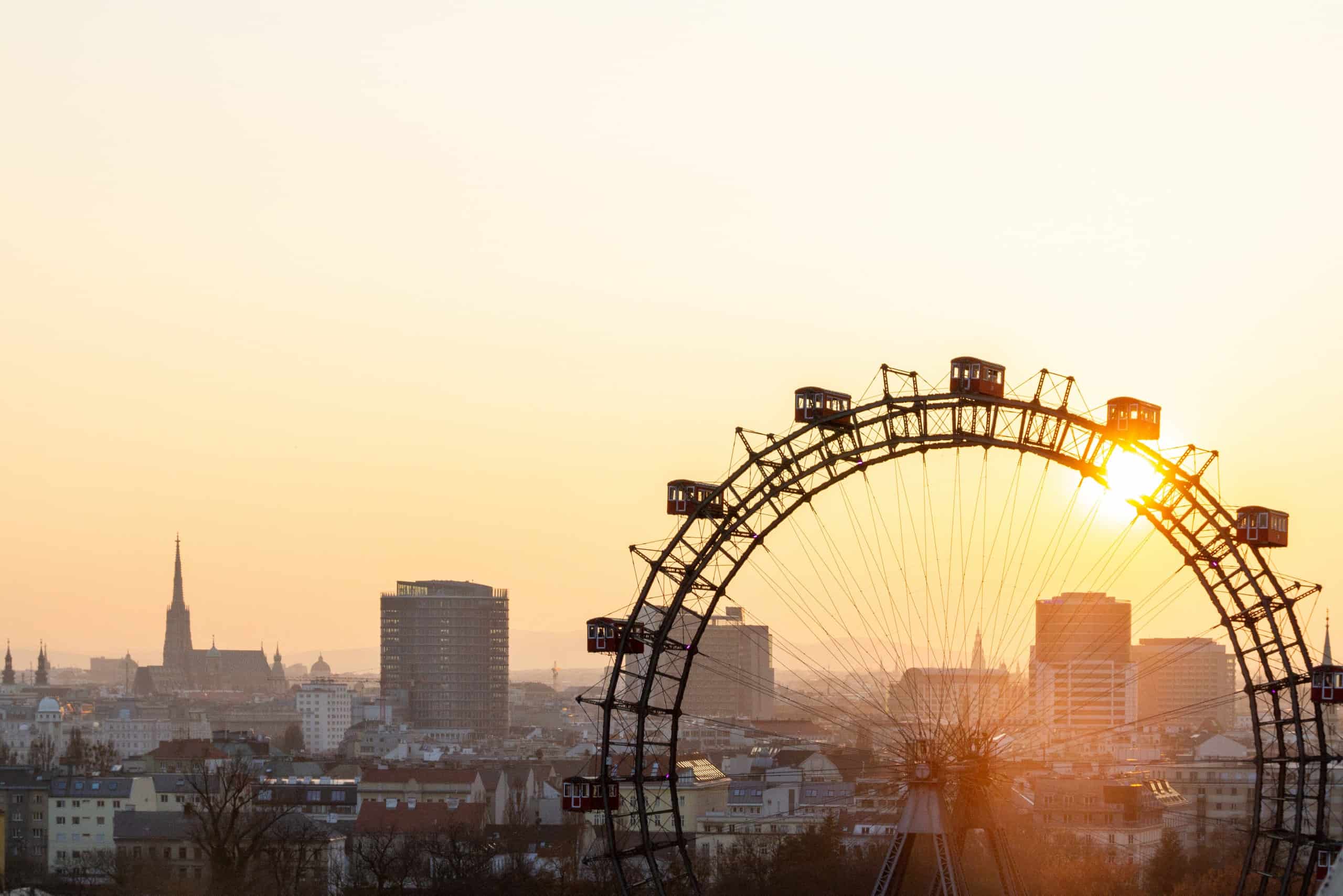 Blick auf das Wiener Riesenrad und die Skyline der Stadt, inklusive Stephansdom, w&auml;hrend die Sonne in warmen T&ouml;nen untergeht