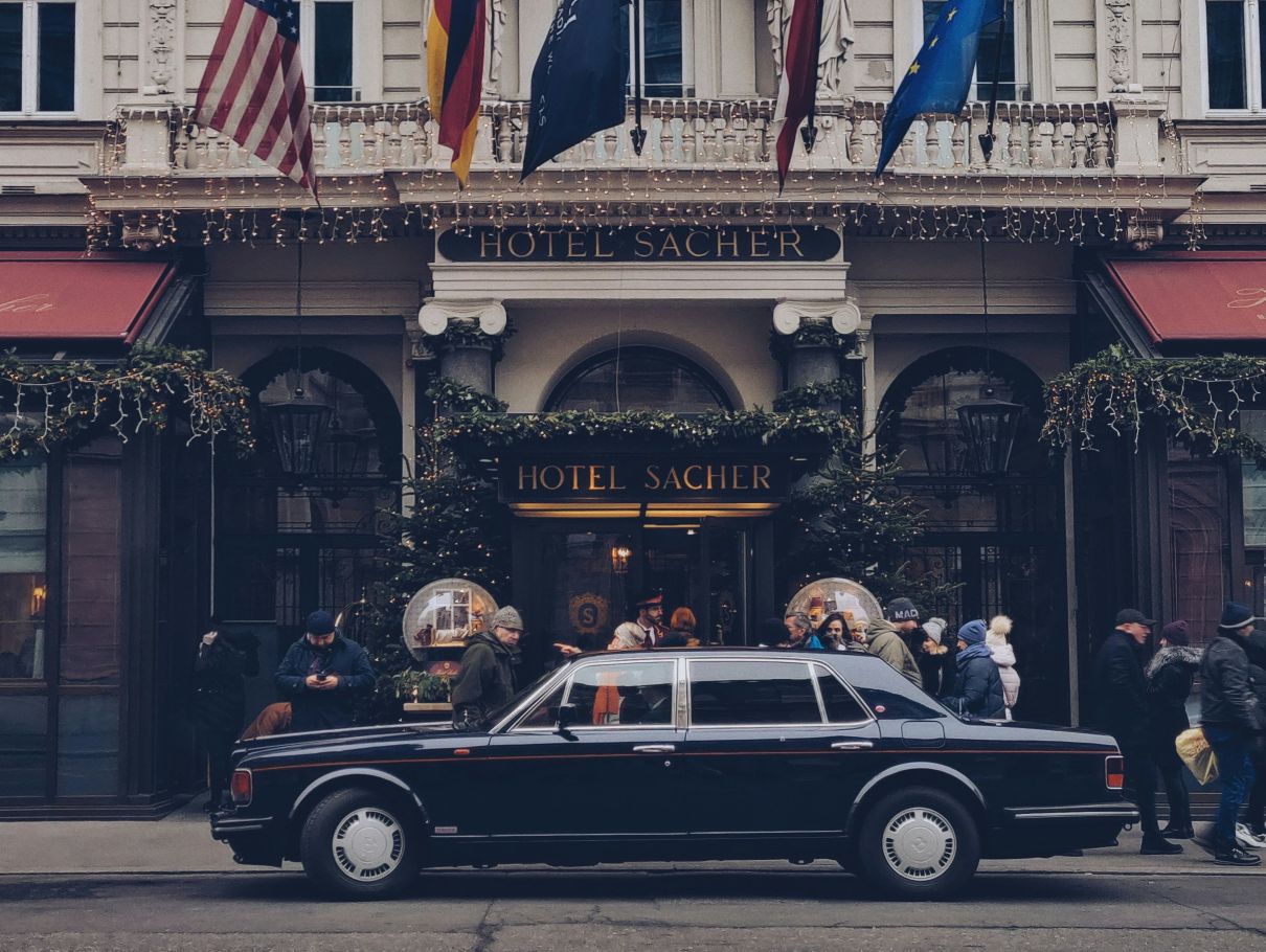 Der elegante Eingang des Hotels Sacher in Wien, geschm&uuml;ckt mit Lichterketten und Fahnen, sowie einem schwarzen Luxusauto im Vordergrund.