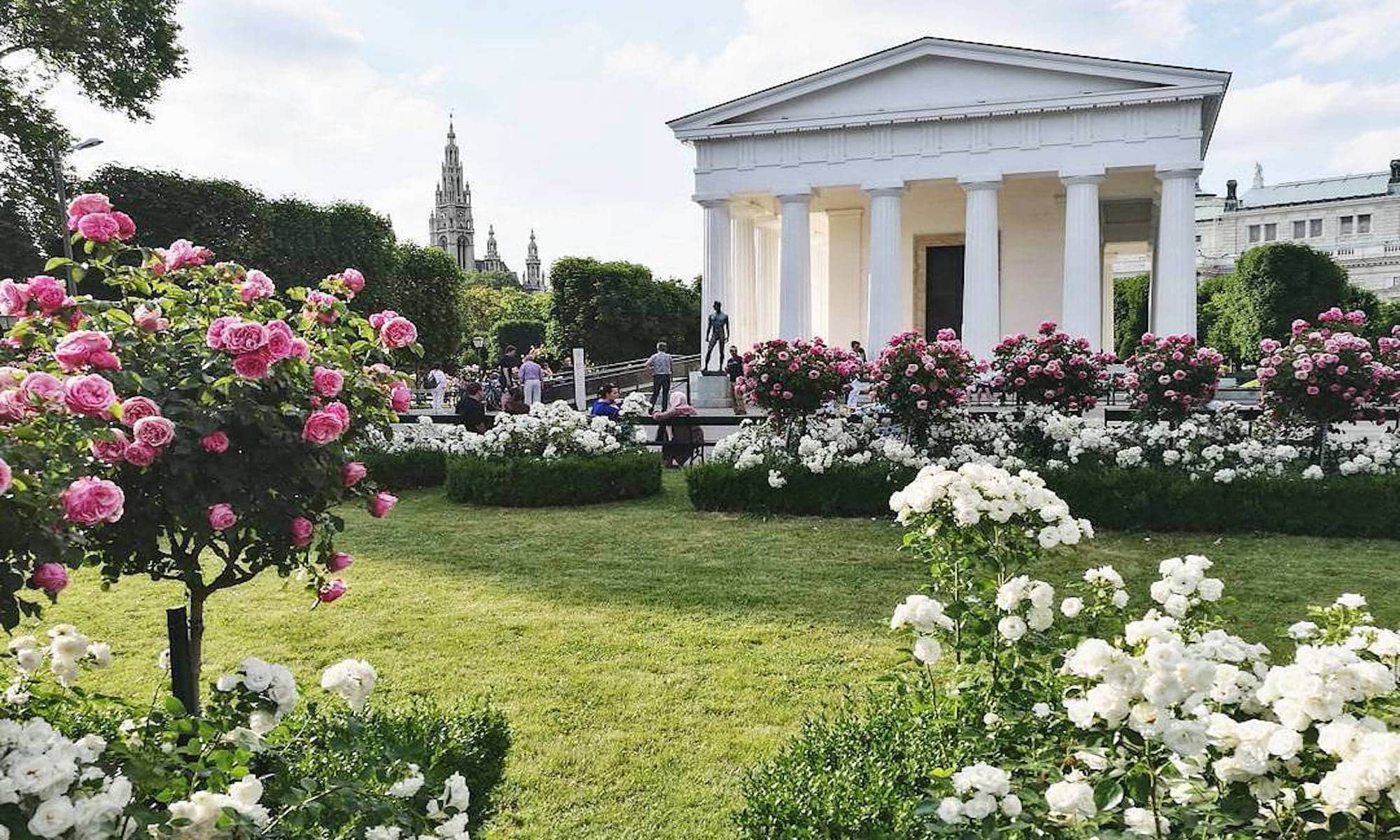 Theseustempel im Wiener Volksgarten umgeben von bl&uuml;henden Rosenbeeten und mit Blick auf die Wiener Architektur im Hintergrund