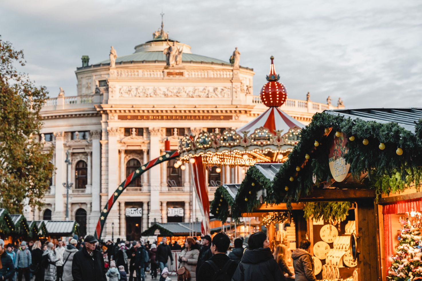 Menschenmengen auf einem festlich geschm&uuml;ckten Christkindlmarkt vor dem Wiener Burgtheater in der D&auml;mmerung.