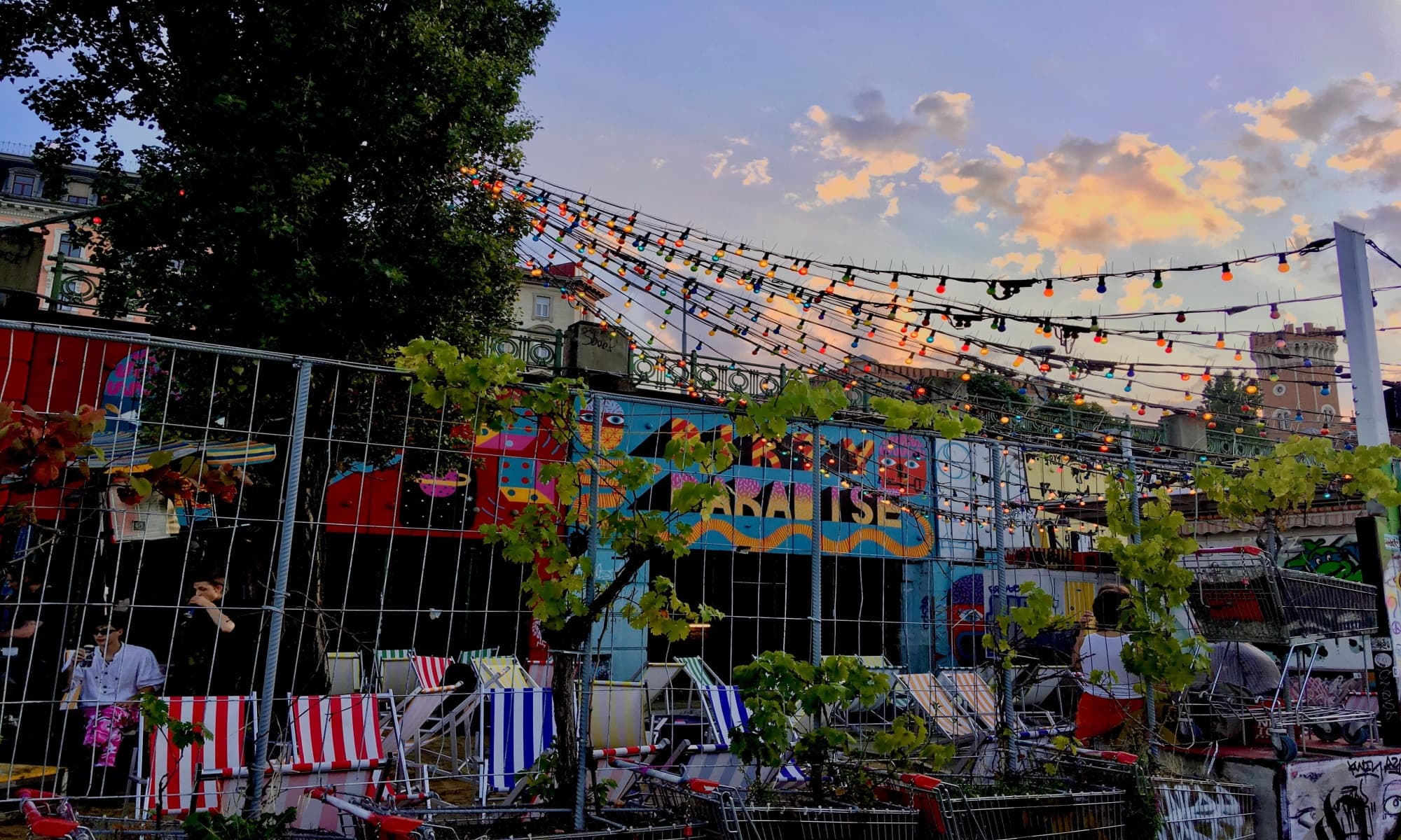 Bunte Outdoor-Bar mit Lichterketten, Streetart, Liegest&uuml;hlen und Einkaufw&auml;gen bei Sonnenuntergang in urbaner Umgebung.