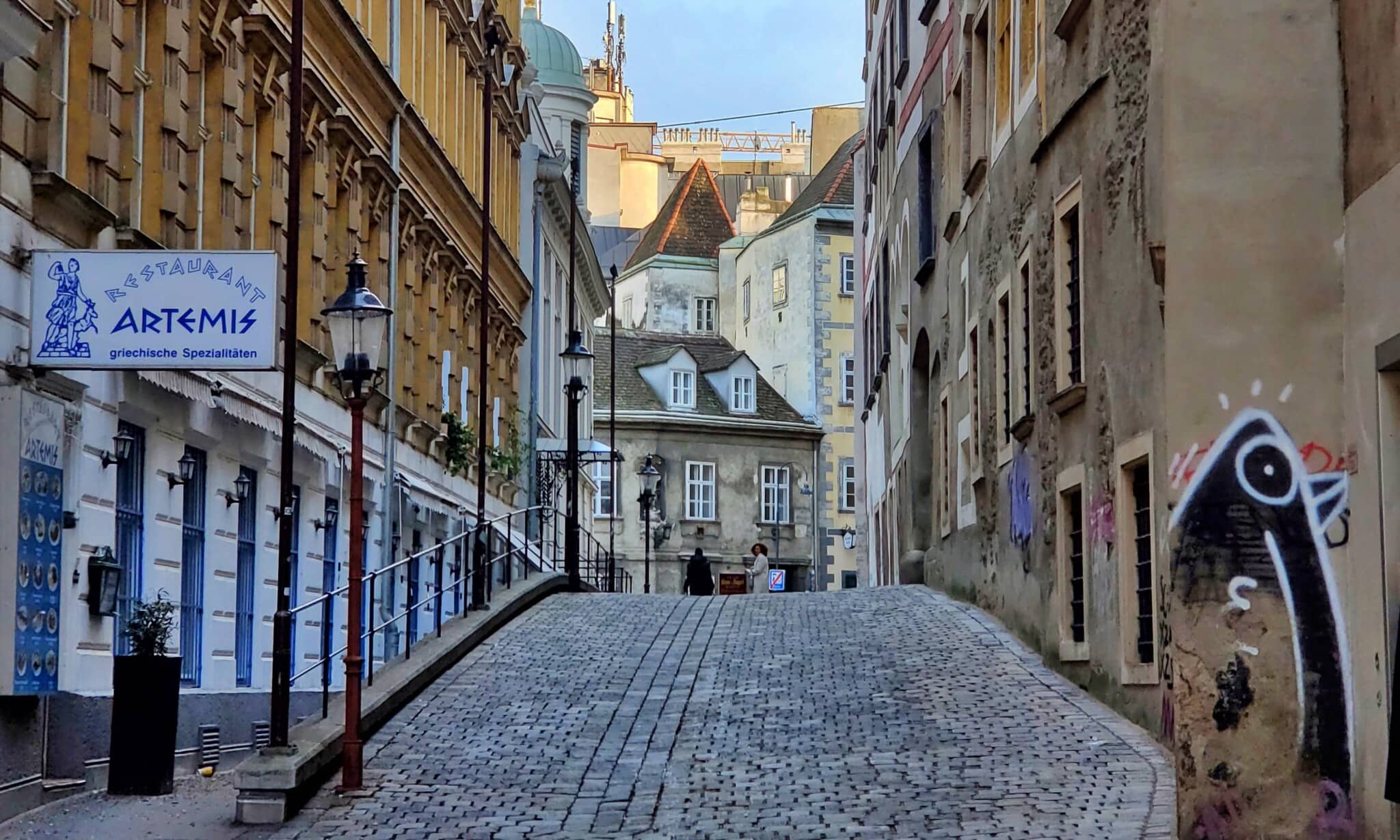 Historische Kopfsteinpflastergasse in Wien mit einem griechischen Restaurant und Graffiti-Kunst an den W&auml;nden, ideal f&uuml;r Stadtrundg&auml;nge.