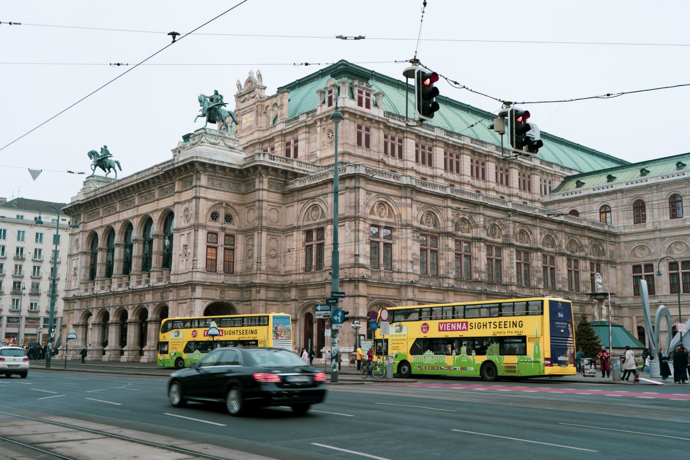 Die Wiener Staatsoper mit gelben Sightseeing-Doppeldeckerbussen im Vordergrund an einer belebten Stra&szlig;e.
