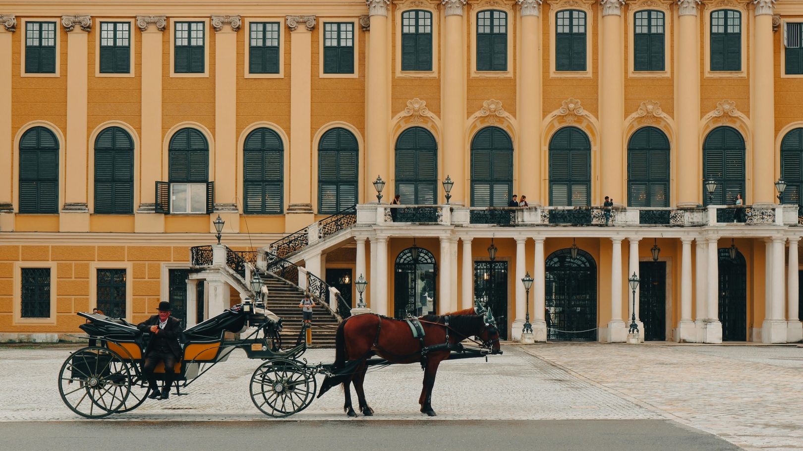 Eine traditionelle Pferdekutsche vor dem Schloss Sch&ouml;nbrunn in Wien, mit historischen gelben Fassaden und Besuchern auf den Balkonen.