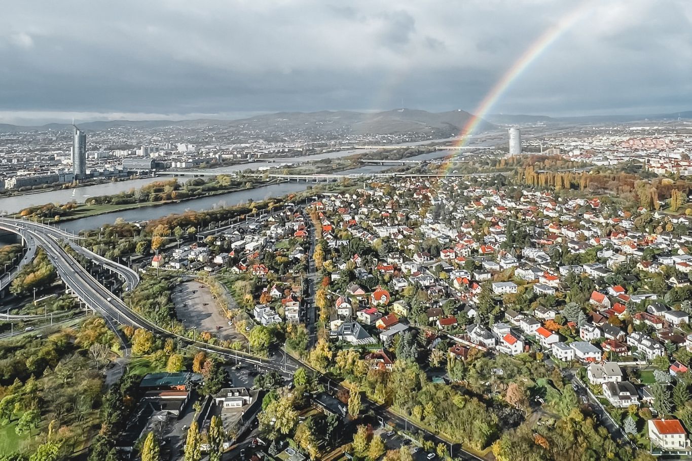 Panoramaaufnahme von Wien mit der Donau, gr&uuml;nen Landschaften und einem farbenfrohen Regenbogen am Horizont