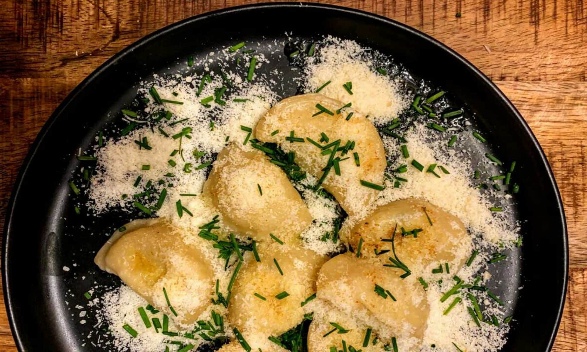 Nahaufnahme eines Tellers mit gef&uuml;llten Teigtaschen, bestreut mit Parmesan und frischem Schnittlauch, serviert auf einem schwarzen Teller
